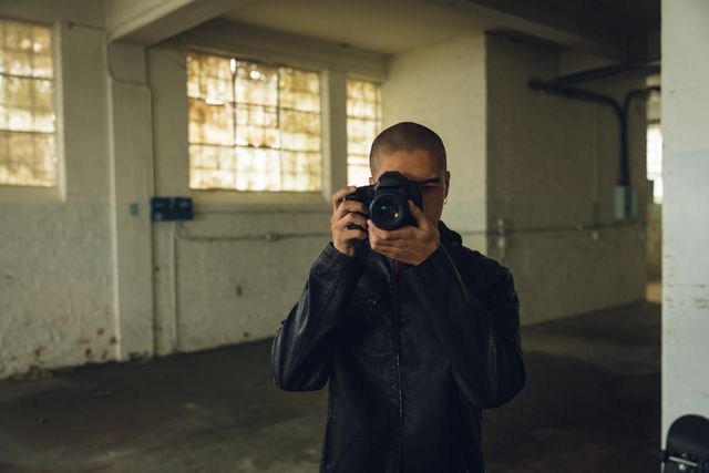 Front view of a hip young biracial man in an empty warehouse, taking photos with SLR camera, wearing black jacked.