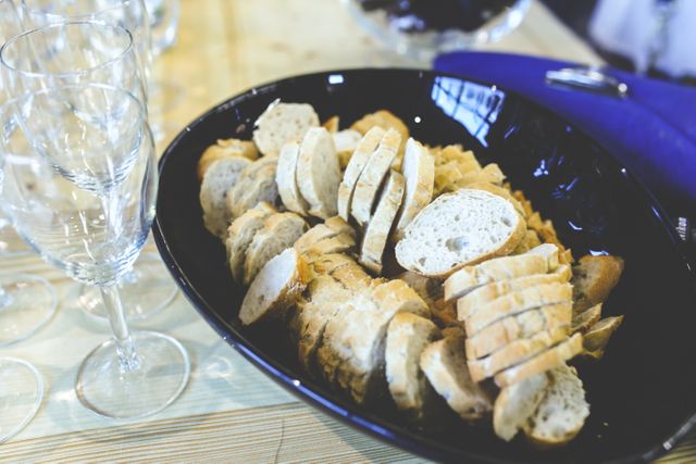 Close-up view of sliced baguette bread neatly arranged in a black bowl beside wine glasses on a dining table. This setup is ideal for illustrating elegant dinner settings, restaurant services, home-cooking blogs, and hospitality events.