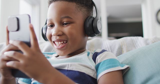 Young boy enjoying time at home, using headphones and smartphone while relaxing on couch. Ideal for topics about childhood, modern technology, leisure time, smart devices, and home comfort.