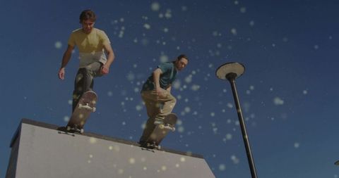 Two skateboarders launching off rooftop ledge urban skate action with streetlamp and bokeh