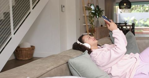 African american woman reclining on sofa listening with headphones and using smartphone