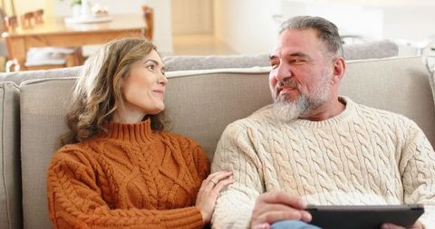 Mature couple sharing cozy moment on sofa wearing cable-knit sweaters and holding tablet