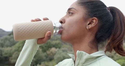 Indian woman drinking from reusable bottle on nature trail promoting hydration and fitness