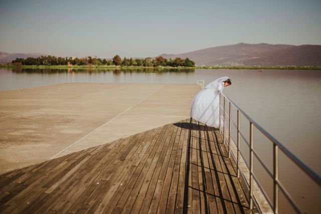 Bride in white gown stands on dock overlooking calm lake on sunny day. Ideal for wedding invitations, bridal magazines, nature photography showcases, romantic scenes, or stock photos depicting calm and serene moments.