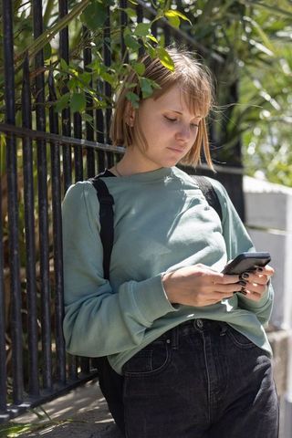 Young woman checking smartphone while leaning on wrought-iron fence on sunny sidewalk