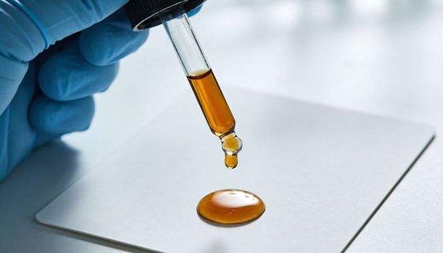 Gloved hand dropping amber liquid from glass dropper onto white laboratory tile
