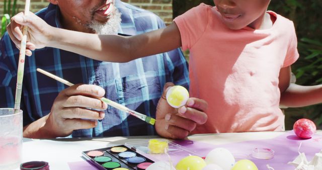 Grandfather engaging in egg painting activity with grandson outdoors on sunny day. They are holding paintbrushes and colorful paints, showcasing moments of bonding and creativity. Ideal for illustrating family activities, intergenerational connections, and creative outdoor pastimes.