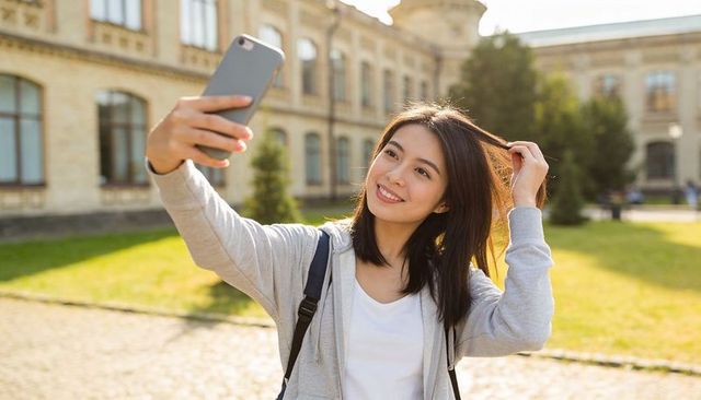 Smiling student taking selfie on sunlit historic campus with backpack and hoodie