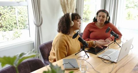 Diverse female cohosts recording podcast at home with microphones, headphones, laptop
