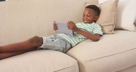 Smiling boy reclining on beige sofa using tablet, relaxed modern living room lifestyle