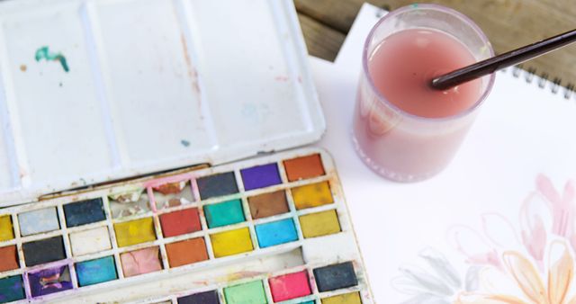 A set of watercolor paints and a brush dipped in water are ready for an artist to create, with copy space. Art supplies like these inspire creativity and are essential for painters to bring their visions to life.
