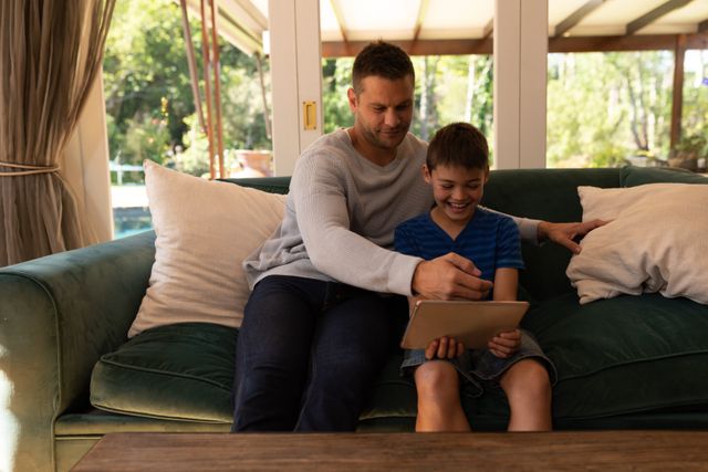 Caucasian father and his son sitting on the couch at their home. the boy is holding a tablet in front of them while smiling.