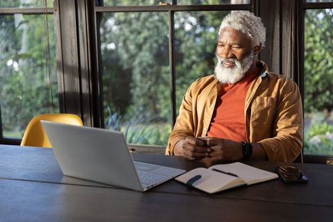 Senior african american man working remotely smiling at laptop in sunlit home office