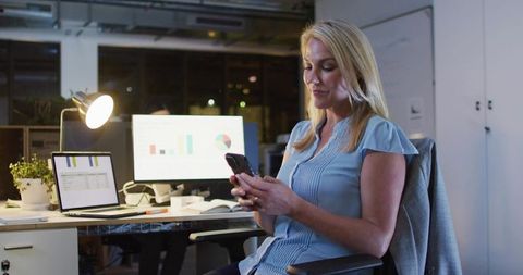 Businesswoman checking smartphone in modern office at night with charts on screens