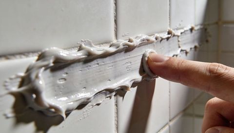 Finger smoothing grey-white sealant along bathroom tile grout for caulking repair and sealing