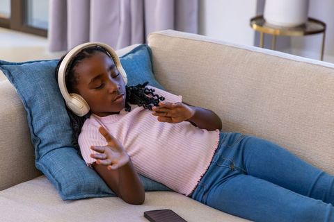 Young african girl listening to music with headphones reclining on sofa at home, relaxing