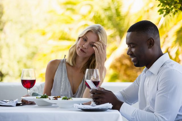 Irritated woman looking at man using phone at table in restaurant