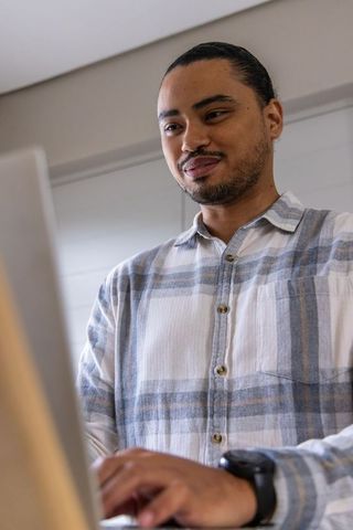 Professional standing at laptop in modern home office wearing plaid shirt and smartwatch