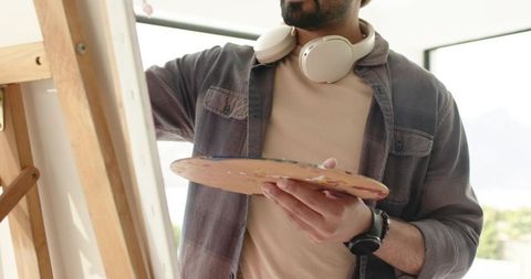 Indian artist painting on canvas at easel in sunlit studio wearing headphones and holding palette