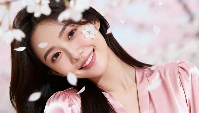 Smiling asian woman tilting head with cherry blossom on cheek wearing satin pink blouse