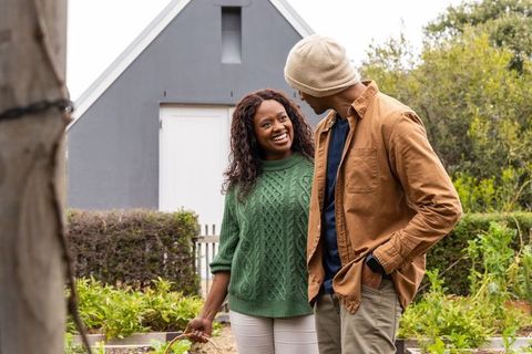 African american couple gardening together by grey shed smiling and holding harvest basket