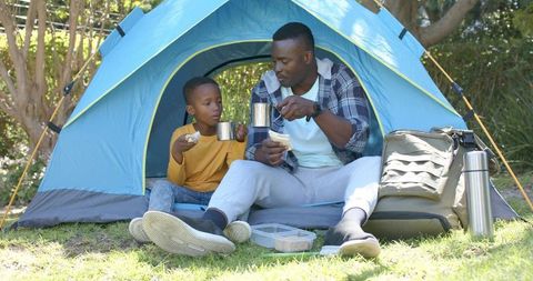 African american father and son camping in blue tent sharing hot drinks and picnic snacks