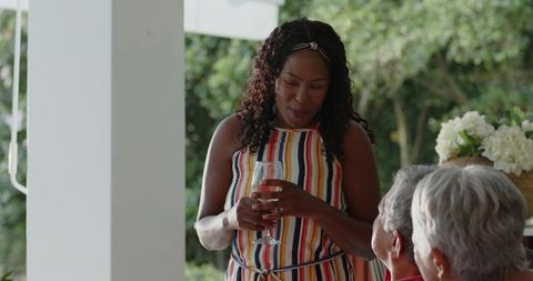 African American woman holding wine glass, chatting with senior couple on sunny porch
