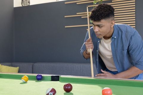 Young african american man leaning over pool table aiming cue in modern lounge