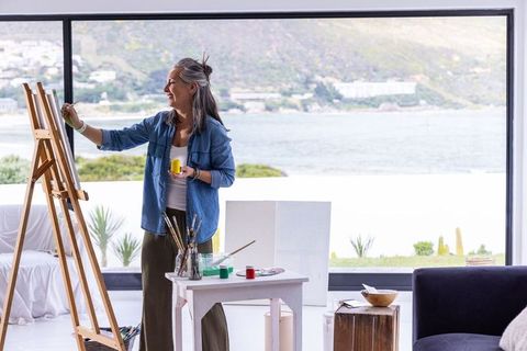 Senior woman painting at easel in coastal home with ocean view, creative studio lifestyle