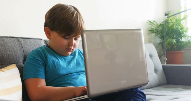 Young boy is sitting on a couch, intently using a laptop for online learning or playing educational games. This image can be used in articles about remote education, homeschooling, child development, and technology. It is also suitable for advertisements for educational software, children's gadgets, and home furniture.