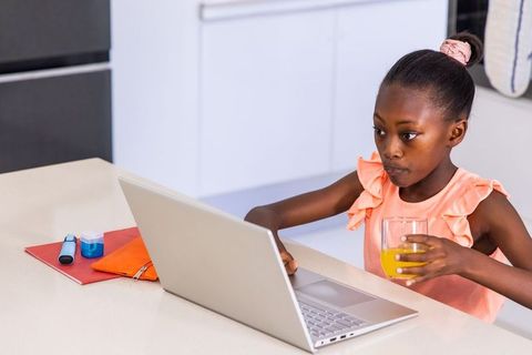 African american girl studying at kitchen counter using laptop and drinking orange juice