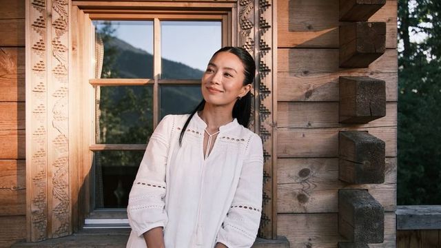 Asian woman relaxing on rustic cabin porch wearing white embroidered blouse, hoop earrings