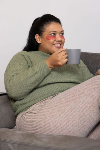 Young woman relaxing on sofa wearing red under-eye patches and holding mug of tea