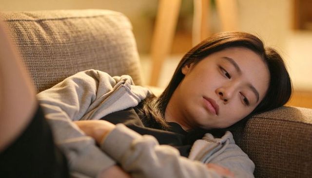 Young woman resting on beige sofa in gray hoodie looking contemplative at home