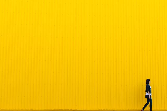 Woman walking alone against a large, vibrant yellow wall. Ideal for use in urban lifestyle, solitude themes, background image for blogs, or animations to depict modern minimalistic design. Perfect for advertising urban fashion or bold color concepts.