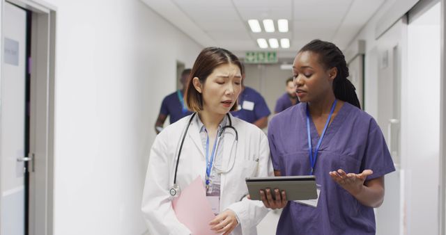 Image of two diverse female doctors walking in busy hospital corridor, looking at tablet and talking. Hospital, medical and healthcare services.