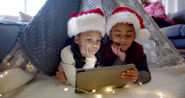 Two children lying in a cozy blanket fort are using a tablet while wearing Christmas hats, emphasizing holiday spirit and technology in a warm, loving family environment. Perfect for themes around childhood joy during celebrations, digital lifestyle, and close sibling relationships. Ideal for family Christmas cards or marketing materials for holiday promotions.