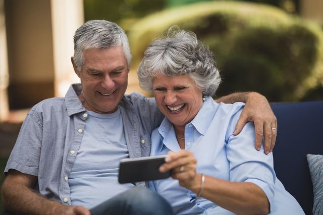 Senior couple sitting on a sofa in their backyard, smiling and looking at a mobile phone. Perfect for themes related to senior lifestyle, technology use among elderly, family bonding, and outdoor leisure activities.