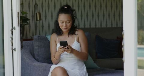 Asian Woman Smiling in White Dress Using Smartphone Indoors