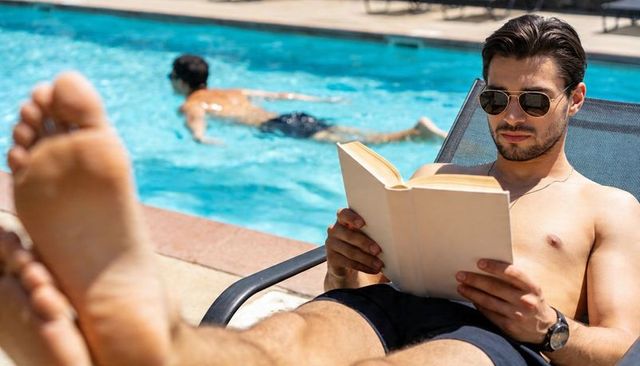 Shirtless man reading on pool lounger wearing aviator sunglasses and swim shorts, sunlit leisure