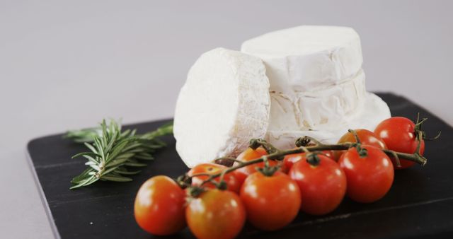 Close-up of a gourmet cheese assortment with ripe cherry tomatoes and fresh rosemary on dark slate. Perfect for promoting culinary delights, restaurant menus, food styling, and gourmet blogs.