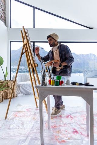 Indian artist painting on easel in minimalist bright studio with mountain and lake view