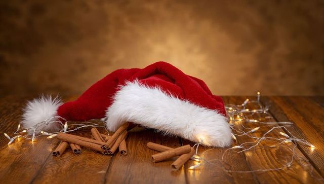 Resting red Santa hat with fur trim on rustic wooden table with cinnamon and fairy lights