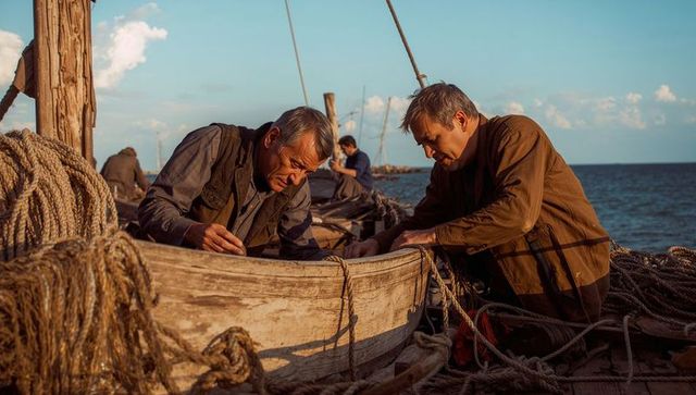 Two fishermen repairing weathered wooden boat on sunlit harbor pier with ropes
