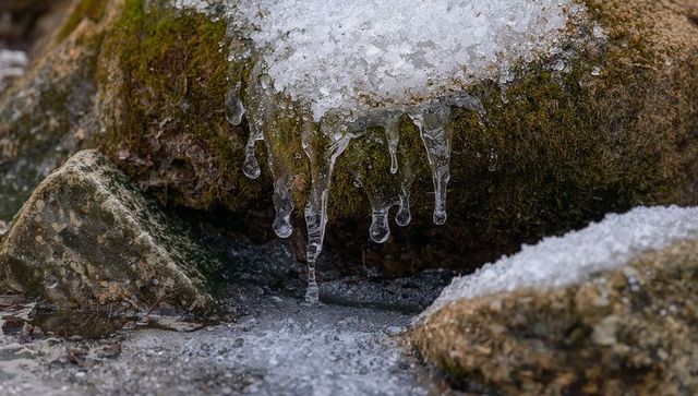 Moss-covered rock dripping icicles over frozen stream bed with granular snow patches
