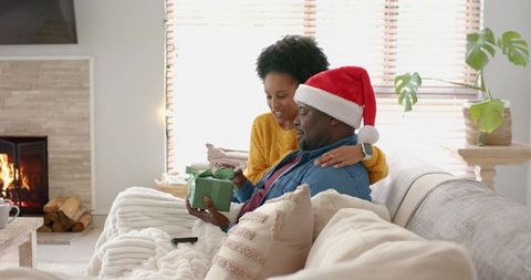 Couple sharing christmas gift on sofa wearing santa hat by cozy fireplace holiday morning