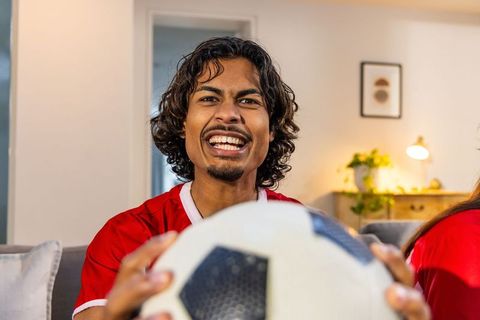 Indian football fan cheering with soccer ball on couch during home match celebration