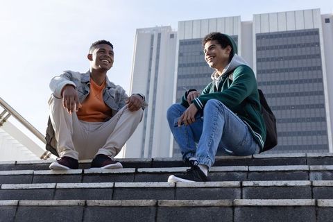Two young men sitting on city steps smiling and chatting with backpacks and highrises