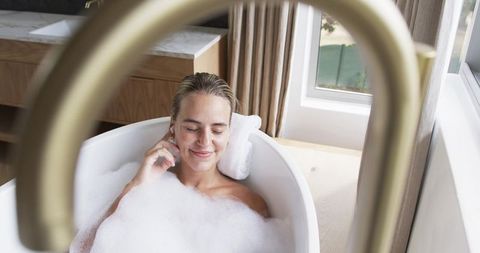 Woman relaxing in freestanding bathtub with bubbling foam and brass faucet, serene spa-style bathroo