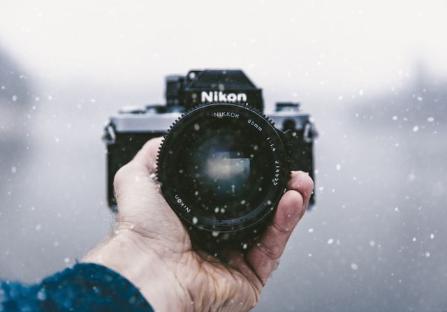 Perfect for illustrating photography hobbies, winter adventures, cold weather gear, technology in nature, or promotional material for camera products and accessories. This detailed shot of a hand holding a film camera in snowy conditions captures both the nostalgia and ruggedness associated with traditional photography.